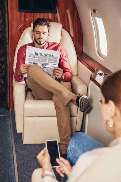 selective focus of boyfriend reading newspaper and girlfriend holding smartphone in private plane  - Photo, Image
