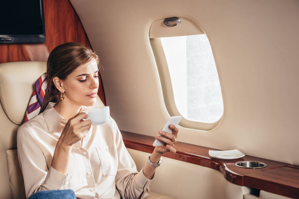 シャツを着た魅力的な女性がカップを持ちプライベート飛行機でスマートフォンを使って  - 写真・画像