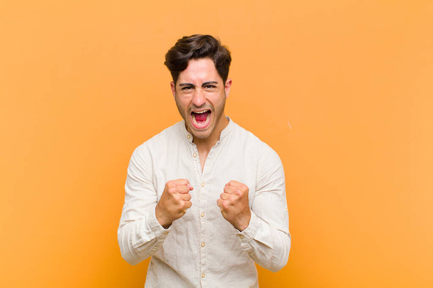 young handsome man shouting aggressively with annoyed, frustrated, angry look and tight fists, feeling furious against orange background - Фото, зображення