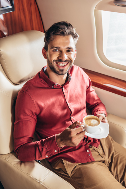 handsome man in shirt smiling and holding cup in private plane  - Фото, изображение