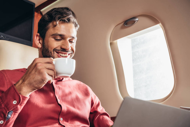 ノートパソコンを使ってシャツを着た笑顔の男とプライベート飛行機でカップを持ち  - 写真・画像