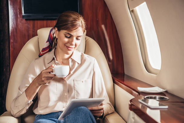 attractive woman in shirt using digital tablet and holding cup of coffee in private plane  - 写真・画像