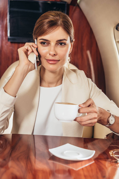 attractive businesswoman in suit holding cup and talking on smartphone in private plane  - Foto, afbeelding