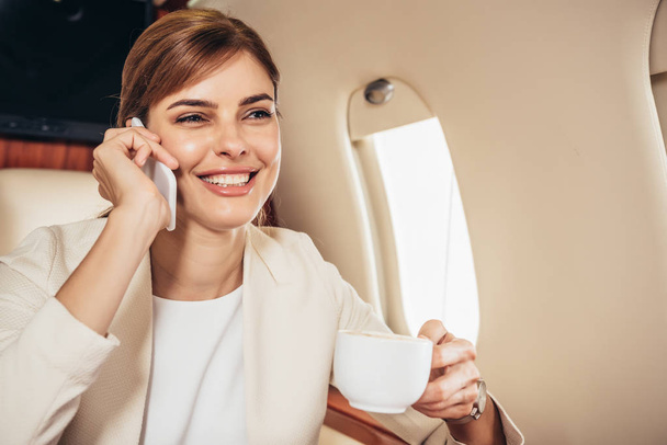 sonriente mujer de negocios en traje sosteniendo taza y hablando en el teléfono inteligente en avión privado
  - Foto, imagen