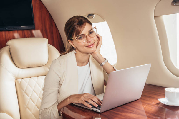 atractiva mujer de negocios en traje con portátil mirando a la cámara en avión privado
  - Foto, imagen