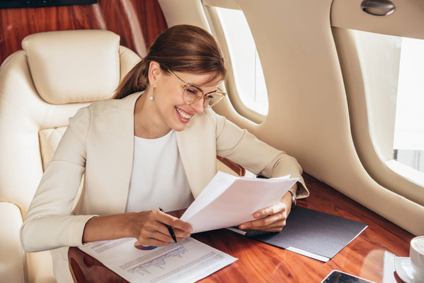 glimlachende zakenvrouw in pak doet papierwerk in prive vliegtuig  - Foto, afbeelding