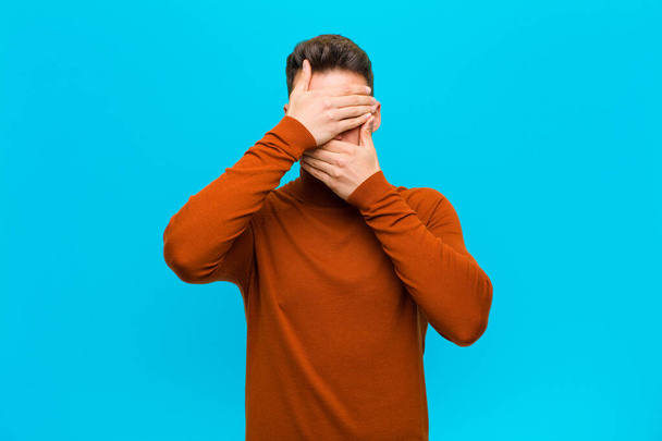 young hispanic man covering face with both hands saying no to the camera! refusing pictures or forbidding photos against blue background - Foto, Imagem