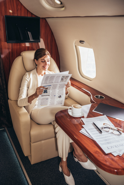 Lächelnde Geschäftsfrau im Anzug liest Zeitung im Privatflugzeug  - Foto, Bild