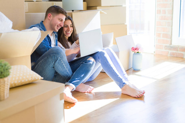 junges Paar sitzt auf dem Boden des neuen Hauses um Kartons herum mit Laptop und lächelt glücklich für neue Wohnung - Foto, Bild