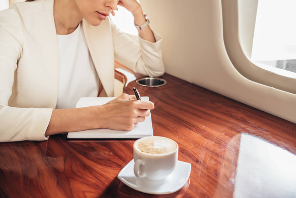 cropped view of businesswoman in suit writing in notebook in private plane  - Foto, Bild