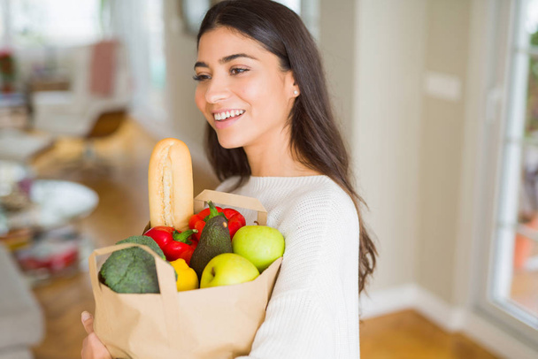 Όμορφη νεαρή γυναίκα χαμογελά κρατώντας μια χάρτινη σακούλα γεμάτη φρέσκα λαχανικά στο σπίτι - Φωτογραφία, εικόνα