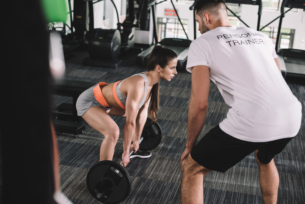 selective focus of personal trainer looking at athletic sportswoman lifting barbell - Photo, Image