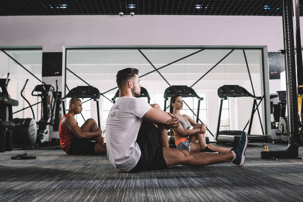 young trainer with multicultural athletes stretching while sitting on floor in gym - Photo, Image