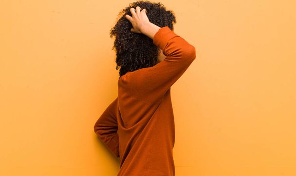 young pretty black woman feeling clueless and confused, thinking a solution, with hand on hip and other on head, rear view against orange wall - Foto, Imagen