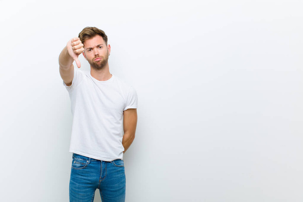 young man feeling cross, angry, annoyed, disappointed or displeased, showing thumbs down with a serious look against white background - Photo, Image