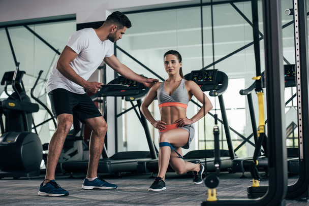 personal trainer holding clipboard while assisting of attractive sportswoman exercising with resistance band - Fotoğraf, Görsel