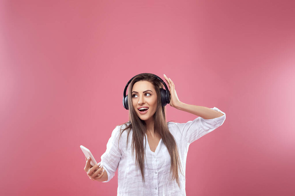Hermosa mujer escuchando música usando auriculares inalámbricos en el estudio aislado sobre fondo rosa. Chica utiliza auriculares inalámbricos y bailando
. - Foto, Imagen