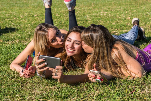 Щасливі друзі в парку в сонячний день. Портрет трьох жінок-друзів насолоджується гарним днем зі своїми смартфонами
. - Фото, зображення