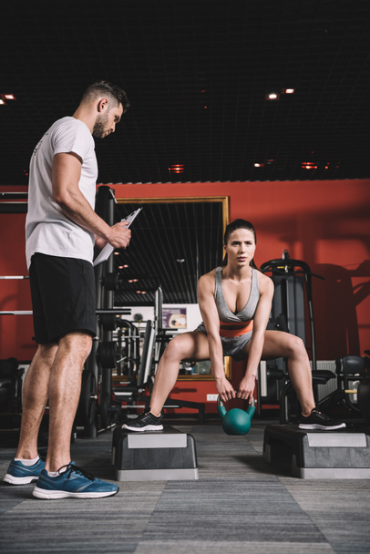 attentive trainer holding clipboard while standing near supervising young sportswoman lifting weight in gym - Photo, Image