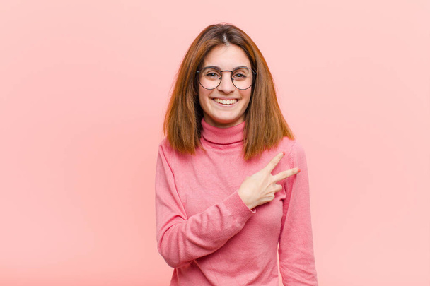 young pretty woman feeling happy, positive and successful, with hand making v shape over chest, showing victory or peace against pink background - Foto, immagini