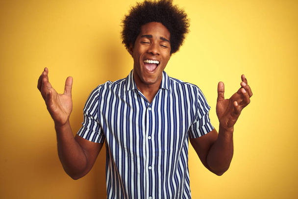 Αμερικανός άνδρας με αφρο μαλλιά φορώντας ριγέ πουκάμισο στέκεται πάνω από απομονωμένο κίτρινο φόντο γιορτάζει τρελός και τρελός για την επιτυχία με τα χέρια υψωμένα και κλειστά μάτια ουρλιάζοντας ενθουσιασμένος. Έννοια νικητή - Φωτογραφία, εικόνα