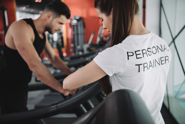 selective focus of personal trainer standing near sportsman running on treadmill - Fotografie, Obrázek