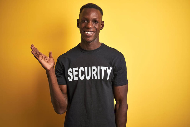 Africká americká ochranná muž nosí bezpečnostní uniformu přes izolované žluté pozadí s úsměvem veselý prezentaci a ukazuje s dlaní ruky při pohledu na fotoaparát. - Fotografie, Obrázek