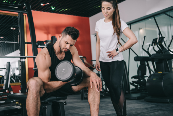 attentive trainer standing near handsome sportsman working out with dumbbell - Photo, Image