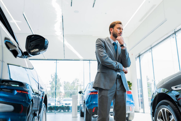 pensive bearded businessman standing near automobiles in car showroom  - Φωτογραφία, εικόνα