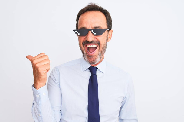 Middelbare leeftijd zakenman dragen schurk leven zonnebril over geïsoleerde witte achtergrond glimlachen met gelukkig gezicht kijken en wijzen naar de zijkant met duim omhoog. - Foto, afbeelding