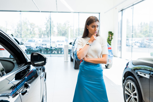 mujer joven y pensativa de pie cerca de los coches en la sala de exposición de coches
  - Foto, imagen