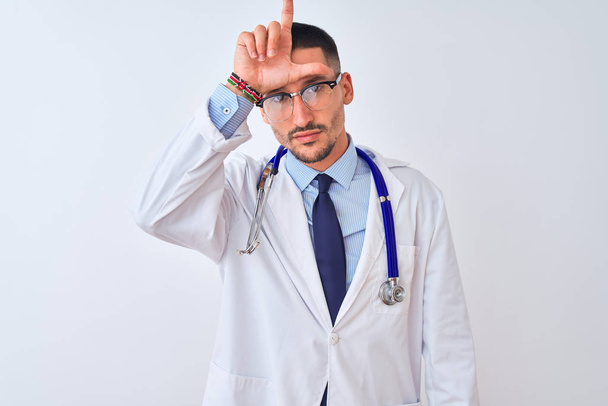 Mladý lékař muž nosit stetoskop přes izolované pozadí dělat si legraci z lidí s prsty na čele dělat poražený gesto posměch a urážlivé. - Fotografie, Obrázek