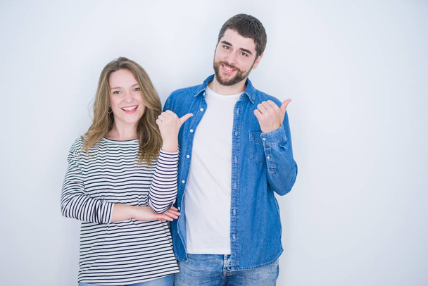 Young beautiful couple standing together over white isolated background smiling with happy face looking and pointing to the side with thumb up. - Fotó, kép