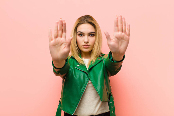 young pretty blonde woman looking serious, unhappy, angry and displeased forbidding entry or saying stop with both open palms against pink flat wall - Fotó, kép