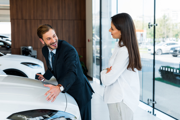 happy bearded car dealer gesturing near attractive woman standing with crossed arms in car showroom  - Fotó, kép