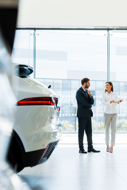 selectieve focus aantrekkelijke auto dealer gebaren in de buurt van bebaarde man in de auto showroom  - Foto, afbeelding