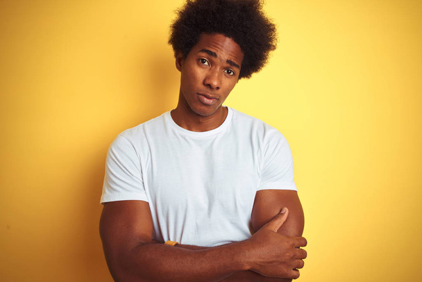American man with afro hair wearing white t-shirt standing over isolated yellow background skeptic and nervous, disapproving expression on face with crossed arms. Negative person. - Photo, Image