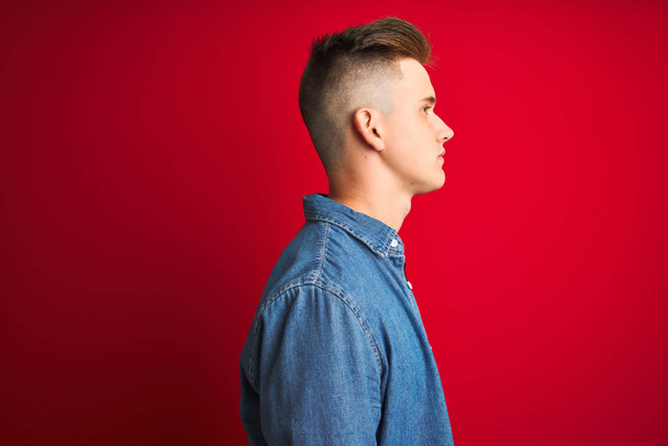 Young handsome man wearing denim shirt standing over isolated red background looking to side, relax profile pose with natural face with confident smile. - Foto, Imagen