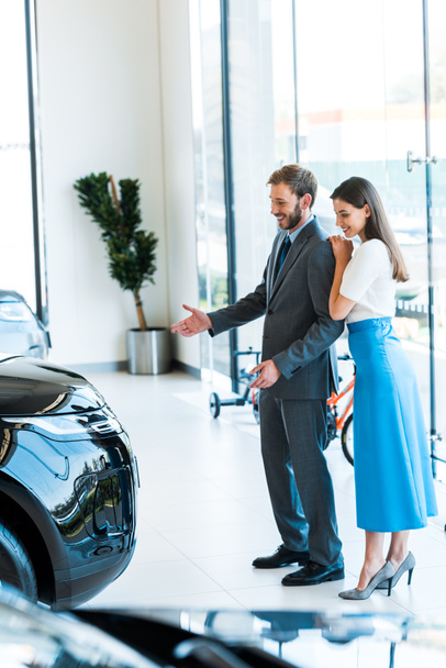 selective focus of cheerful woman standing with handsome bearded man gesturing while looking at car  - Photo, Image