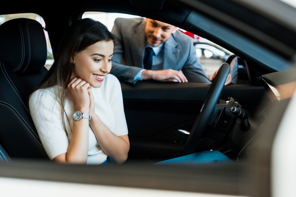 selektywna ostrość uśmiechnięta dziewczyna siedzi w samochodzie w pobliżu brodatego mężczyzny w salonie samochodowym  - Zdjęcie, obraz