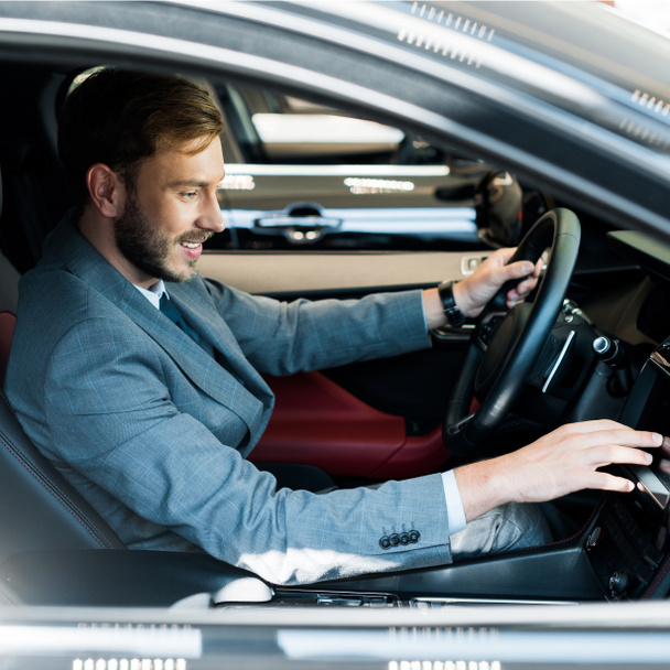 selective focus of cheerful bearded driver sitting on car and touching display  - 写真・画像