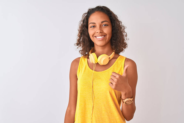 Mujer brasileña joven con auriculares amarillos sobre fondo blanco aislado haciendo pulgares felices gesto hacia arriba con la mano. Aprobar la expresión mirando a la cámara con éxito
. - Foto, Imagen