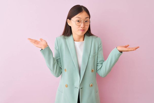 Giovane donna d'affari cinese indossa giacca e occhiali su sfondo rosa isolato espressione indiscussa e confusa con braccia e mani sollevate. Concetto di dubbio
. - Foto, immagini