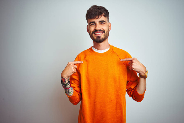 Młody człowiek z tatuażem w pomarańczowym swetrze stojący nad odizolowanym białym tłem z uśmiechem na twarzy, wytykający się palcami dumną i szczęśliwą. - Zdjęcie, obraz