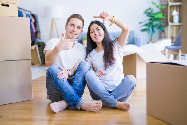 Молодая красивая пара сидит на полу в новом доме вокруг картонных коробок улыбаясь делая рамку с руками и пальцами с счастливым лицом. Концепция творчества и фотографии
. - Фото, изображение