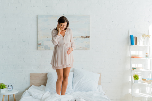 sonriente mujer embarazada tocando su vientre mientras está de pie en la cama en casa
  - Foto, Imagen