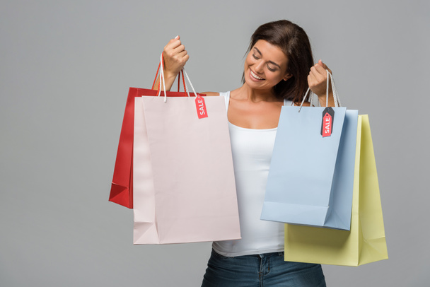 χαρούμενη γυναίκα που κρατάει τσάντες με πινακίδες πώλησης, απομονωμένη σε γκρι - Φωτογραφία, εικόνα