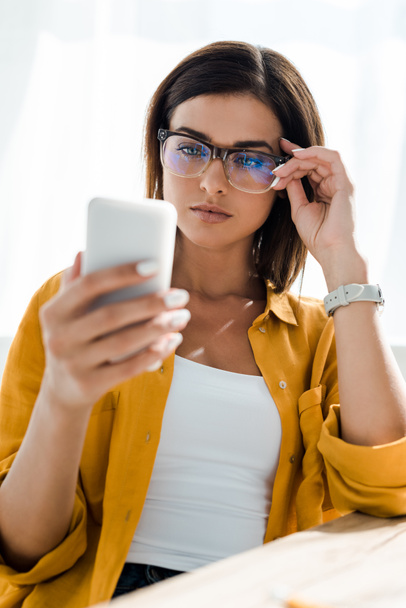 ελκυστική αυτοπεποίθηση freelancer σε γυαλιά με χρήση smartphone στο γραφείο στο σπίτι - Φωτογραφία, εικόνα