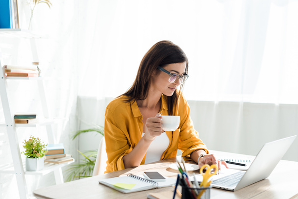 mooie freelancer werken aan laptop met koffiekop in huis kantoor - Foto, afbeelding