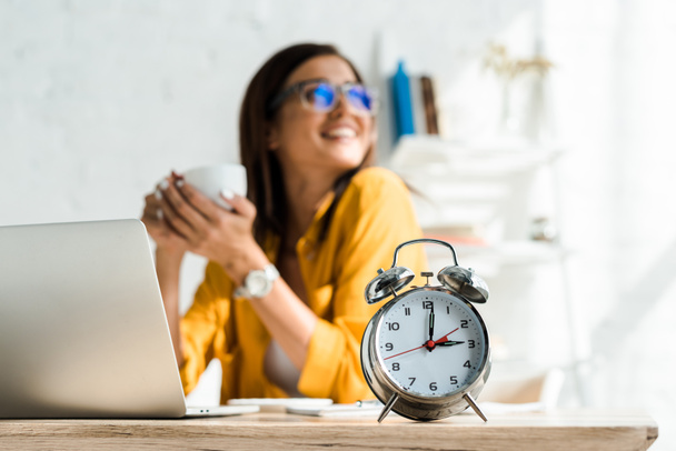 избирательный фокус будильника и счастливый фрилансер с кофе перерыв в домашнем офисе с ноутбуком
 - Фото, изображение